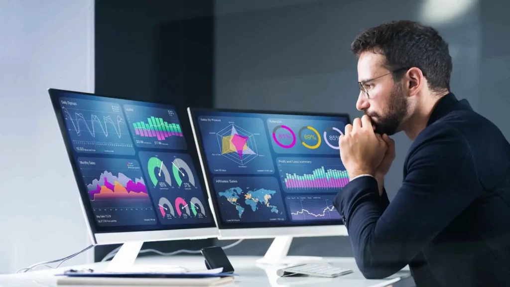 Homem com roupa preta e óculos, apoiando em uma mesa com dois computadores grandes exibindo dados e gráficos, sugerindo análise de dados.