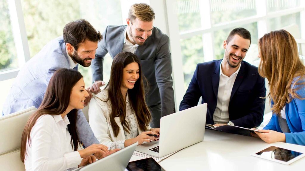 grupo de profissionais sorrindo e colaborando em reunião com laptops e documentos.