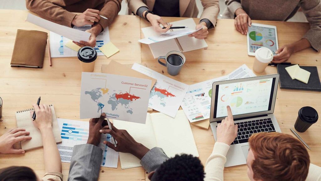 pessoas reunidas em mesa de trabalho analisando gráficos, mapas e relatórios com laptops e tablets.